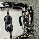Detail bílého bubnu s chromovaným obručem a kování. Na korpusu je připevněno chromované držadlo a mechanismus s knoflíkem. Povrch kování je lesklý, b…