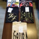 Pět triček s potisky hudebních kapel rozložených na hnědém stole. Jsou zde dvě bílá a tři černá trička s nápisy FOO FIGHTERS, Foo Fighters a GOJIRA.…