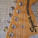 Hlava elektrické kytary z přírodního dřeva s chromovanými ladičkami. Na povrchu je černý nápis Squier STRATOCASTER a pod ním menší text BY FENDER. Po…