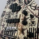 Detail těla elektrické kytary s bílým povrchem a černobílým grafickým potiskem obsahujícím texty, hvězdy a symboly. Nástroj má dva černý humbuckery s…