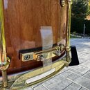 Detailní záběr na část bubnu s hnědým dřevěným povrchem a zlatými kovovými komponenty. Na korpusu je připevněný kovový mechanismus s černou gumovou p…