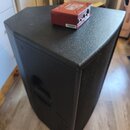 Černý reproduktor s texturovaným povrchem a integrovanou rukojetí na boku. Na horní části reproduktoru leží malý červený kovový box s nápisem MCD a n…
