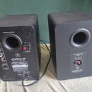 Dva černé studiové monitory s matným povrchem. Na levém je nápis POWERED SPEAKER a vstupy označené jako INPUTS, na pravém nápis PASSIVE SPEAKER INPUT…