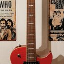 Červená elektrická kytara s tvarem připomínajícím Les Paul, umístěná na nástěnním držáku. Na hlavě je bílý nápis Washburn a logo ve tvaru stylizované…