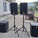 Sada čtyř černých reproduktorů značky JBL. Dva menší reproduktory jsou umístěny na černých tripodních stojanech a dva větší subwoofery stojí na zemi.…