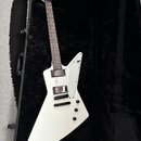 Bílá elektrická kytara s ostrou hranatou formou těla, položená v černém tvrdém kufru. Na hmatníku jsou dva humbuckery a tři ovládací knoflíky, tělo j…