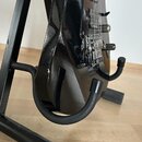 Černá elektrická kytara s lesklým povrchem umístěná na černém stojanu. Na těle kytary jsou vidět dva humbuckery, ladicí kolíky a ovládací prvky. Na n…