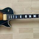 Elektrická kytara s tónem sunburst a černým povrchem na okrajích. Na hlavě je logo Gibson a nápis Les Paul Custom. Nástroj má zlatý hardware, dva hum…