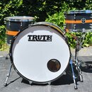 Černá bubnová sada s oranžovými vodorovnými pruhy a chromovaným kováním. Na bílém předním obalu basového bubnu je černý nápis TRUTH DRUMS. Na tomu, k…