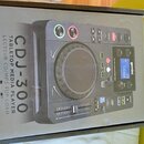 Krabice s obrázkem černého DJského přehrávače. Na obalu je uveden text „gemini DJ“, „CDJ-300 TABLETOP MEDIA PLAYER LECTEUR CD/MP3 TOP LOAD“ a na disp…