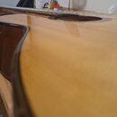 Detailní pohled na tělo akustické kytary s přírodním dřevěným povrchem. Je vidět část hradla, pražců a zvukový otvor. Nástroj nemá žádné viditelné zn…