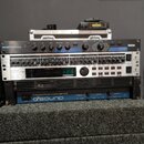 Sada rackového audio vybavení v černém case. Z horní části dolů jsou vidět zařízení s nápisy: Lexicon MPX 100 Dual Channel Processor, nečitelný příst…