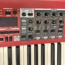 Červený digitální klavír s ovládacím panelem obsahujícím různé přepínače, knoflíky a displej. Na panelu jsou viditelné nápisy sekcí PIANO, PROGRAM, S…