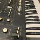 Detailní pohled na ovládací panel a klávesnici elektronického nástroje s černým povrchem. Na panelu jsou umístěny posuvníky, otočné knoflíky a přepín…