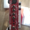 Zadávací panel červeného elektronického zařízení s různými konektory. Jsou zde nápisy MIC IN, INST IN (A/MONO, B), OUTPUT (A/MONO, B), OUT, MIDI IN a…