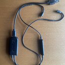 Černý kabel s USB konektorem na jednom konci a dvěma dalšími koncovými plugy. Na kabelu jsou dva černé oválné moduly, z nichž jeden nese svislý nápis…