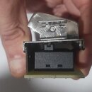 Detailní záběr na kovovou součástku, pravděpodobně konektor nebo část šasi elektronického zařízení. Součást je stříbrná, s viditelným šroubem a dvěma…
