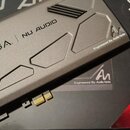 Šedá zvuková karta s kovovým krytem, na kterém je nápis EVGA | NU AUDIO a logo Engineered By Audio Note. Na povrchu jsou vyfrézované geometrické tvar…