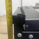 Detailní záběr na hranu černého kovového zařízení, vedle kterého je přiložena žlutá skladovací metr. Na horní hraně je viditelný černý textilní nebo…