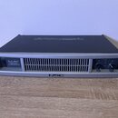 Stříbrný rackový výkonový zesilovač s černým horním krytem. Na předním panelu je nápis PLX 1804 PROFESSIONAL POWER AMPLIFIER a v dolní části logo QSC…