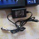 Černý síťový adaptér s kabelem a konektory. Na boku adaptéru je nápis AC/DC ADAPTOR a značka CE. V horní části fotky je vidět část modrého elektronic…