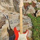 Červená elektrická kytara typu Stratocaster s bílým pickguardem a světlou hnedou hmatníkovou deskou. Na hlavě kytary je viditelné logo Fender. Nástro…