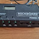 Černý napájecí zdroj Rockboard Power Pit by WARWICK. Na přední straně je LCD displej, přepínač napětí (9V, 12V, 15V, 18V) a nápis „Unplug pedal when…