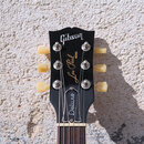 Černá hlava elektrické kytary s nápisy Gibson, Les Paul MODEL a Deluxe. Nástroj má šest ladiček s krémovými knoflíky. Povrch je hladký a bez viditeln…