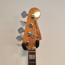 Záběr na hlavu basové kytary z přírodního dřeva s čtyřmi chromovanými ladičkami. Na zadní straně hlavy je černý nápis Fender Jazz BASS a pod ním séri…
