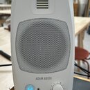 Bílý studiový monitor značky ADAM AUDIO s kovovou mřížkou reproduktoru. V dolní části jsou umístěny ovládací prvky: podsvícené tlačítko s ikonou, vst…
