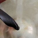 Detailní záběr na povrch kovového činely s koncentrickými rýhami. V horní části je vidět nečitelný nápis. Přes střed činely je položen černý kožený p…