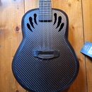 Černý ukulele s tělem z karbonového vlákna v pletené struktuře. Nástroj má čtyři struny a dvě ozdobné otvory v těle ve tvaru stylizovaných plamenů. N…
