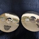 Dvě kovové činely v zlatavé barvě s kovaným povrchem, položené na tmavě modré látce. Levý činele má nápis SABIAN a pod ním grafický symbol v podobě p…