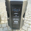 Černý aktivní reproduktor s plastovým povrchem. Na zadním panelu je nápis THUMP 12 a text 1000W 12" POWERED LOUDSPEAKER. Panel obsahuje dva modré oto…