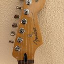 Záběr na hlavu elektrické kytary z přírodního dřeva s šesti chromovanými ladičkami. Na povrchu je černý nápis Fender a STRATOCASTER®, v horní části j…