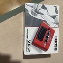 Červený vokální procesor Boss VE-2 Vocal Harmonist umístěný na původním kartonovém obalu. Na zařízení je vidět displej, několik ovládacích knoflíků a…