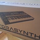 Kartonová krabice s černým potiskem syntetizátoru. Na krabici jsou nápisy HYDRASYNTH, DIGITAL WAVE MORPHING SYNTHESIZEDER a webová adresa www.AshunSo…