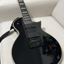 Černá elektrická kytara s tvarem typu Les Paul a bílým lemováním korpusu. Má dva humbuckery, čtyři ovládací knoflíky a černý popruh. Na hmatníku jsou…