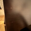 Detailní záběr na černý, matný povrch nábytku nebo části nástroje. Na povrchu jsou viditelné dvě drobné rýhy a několik jemných škrábanců. Žádná značk…
