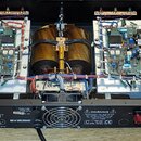 Fotografie zachycuje otevřený vnitřek elektronického zařízení, pravděpodobně zesilovače, s dvěma identickými moduly obvodů a centrálním transformátor…