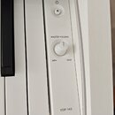Detail ovládacího panelu bílého digitálního piana. Panel obsahuje tlačítko napájení s ikonou zapnutí, otočný knoflík s nápisy MASTER VOLUME, MIN a MA…