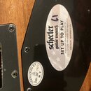Detail zadní strany kytary s černým plastovým krytem a dřevěným tělem. Na krytu je oválná nálepka s nápisy Schecter guitar research 61, SET UP TO PLA…