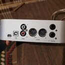 Stříbrný kovový přístroj s řadou konektorů na zadní straně. Jsou zde nápisy +5V DC, USB, S/PDIF (s vstupy In a Out), midi I/O (s konektory Out a In)…