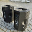 Dvě černé aktivní reproduktory s texturovaným povrchem umístěné na dlažební kostce. Na zadním panelu jsou viditelné ovládací prvky, konektory a nápis…