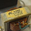 Fotografie zachycuje elektrický transformátor s kovovým jádrem a měděným vinutím. Na horní straně je žlutá nálepka s nápisy: A-2905, MERIT (vedle log…