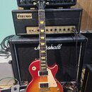 Elektrická kytara s tvarem singlecut a povrchovou úpravou sunburst v červeně a žluté. Na hlavě je bílé logo Gibson a nápis Les Paul Custom. Kytara má…