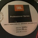 Fotografie detailu štítku na zadní straně reproduktoru. Na černém pozadí je oranžové logo JBL a nápis Professional Series. Dole jsou uvedeny texty JA…