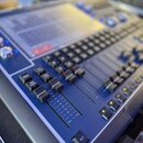 Modrá digitální mixovací konzole s displejem a řadou posuvníků. Na ovládacích prvcích jsou viditelné nápisy "DBS", "ADD", "SWAP" a "ALT". Povrch přís…