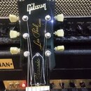 Černá hlava elektrické kytary s bílým logem Gibson. Na povrchu je zlatý nápis Les Paul CLASSIC a níže v dekorativním rámu nápis Classic. Kytara má še…
