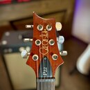 Detail hlavy hnedkové elektro kytary s šesti ladicími mechanismy. Na povrchu je zlatý nápis v kurzívě „Paul Reed Smith“ a na černé vložce u hрифu je…